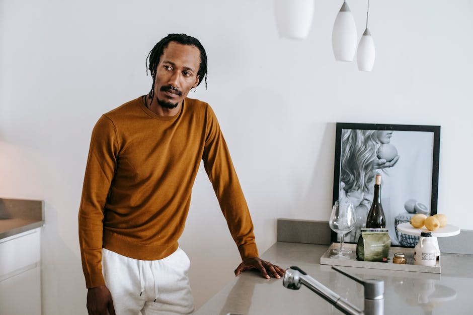 Young African American man leaning on counter at kitchen while looking away and wearing casual clothes in daylight