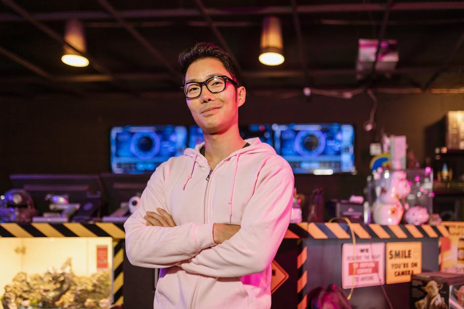 Asian man in pink hoodie confidently standing in an e-sports arena with crossed arms, smiling.
