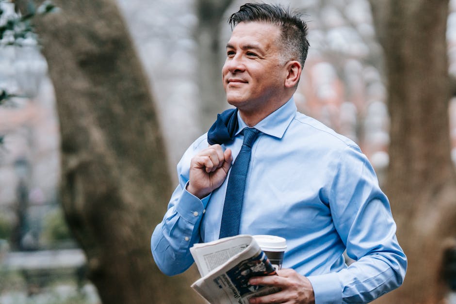 Smiling businessman in a blue shirt, holding coffee and newspaper outdoors.