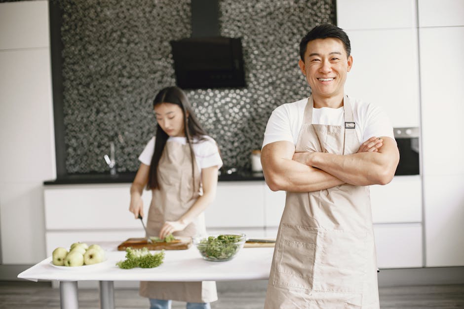 Smiling chef in apron stands confidently in a modern kitchen with a woman preparing food.