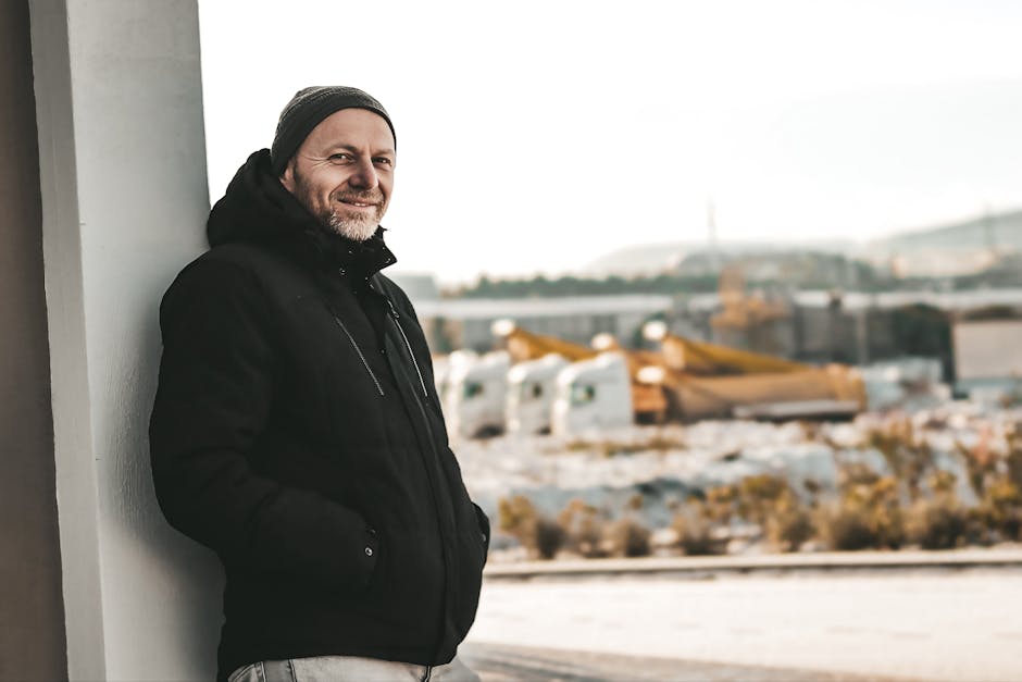 Smiling man in winter attire outdoors leaning against a wall with snowy background.