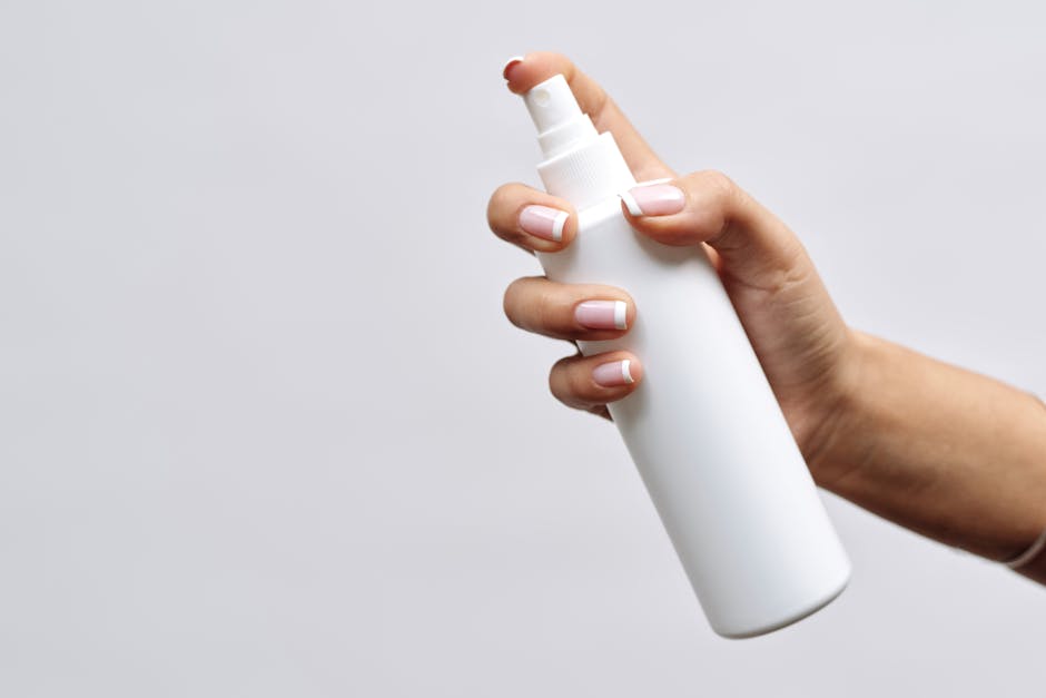 A hand with manicured nails holds a minimal white spray bottle on a white background, perfect for branding mockups.