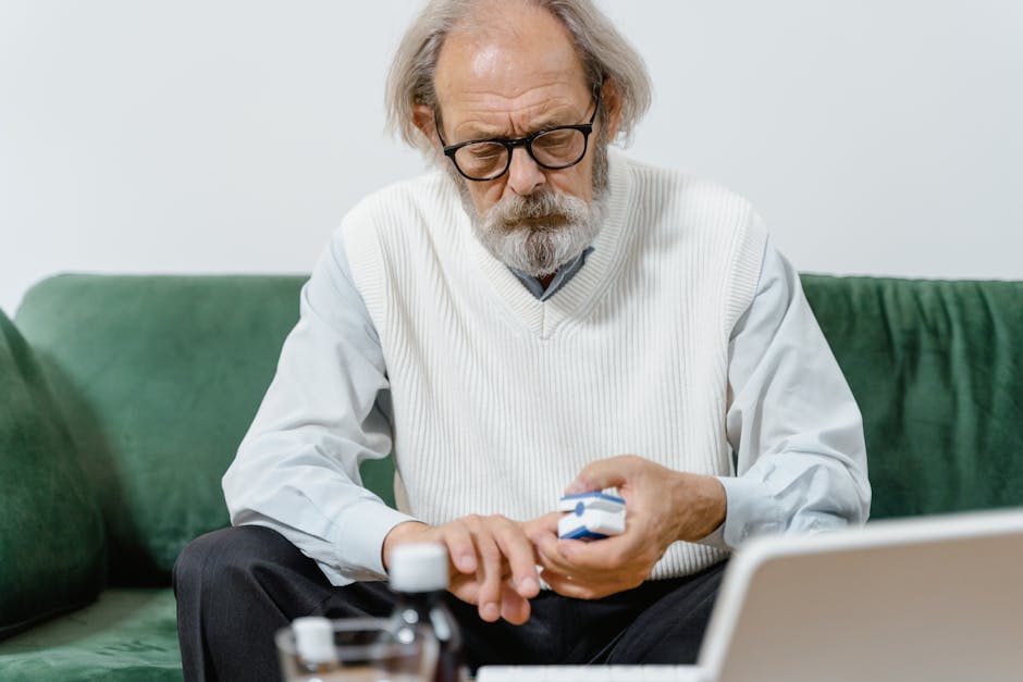Elderly man with eyeglasses carefully examining his medication while seated on a green couch.